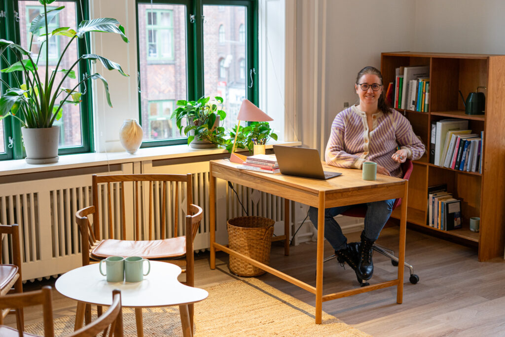 Psykolog sidder klar til at give online psykologhjælp i et roligt rum, kigger venligt ind i kameraet og smiler, med en bærbar computer foran sig.